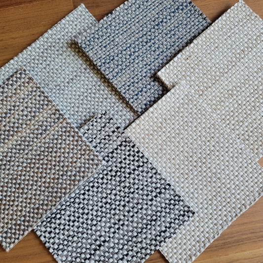 Six woven outdoor rug swatches in different colors and patterns, arranged diagonally on a wooden floor. The swatches include various combinations of beige, gray, white, and blue.